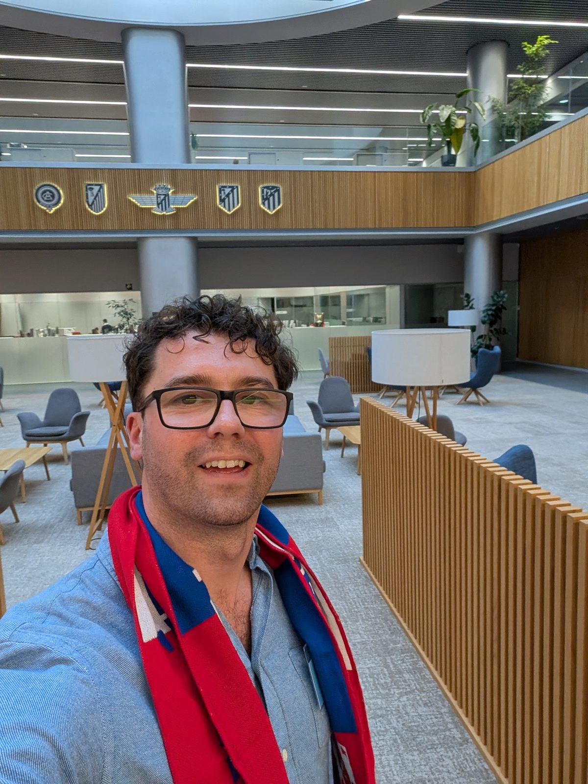 A selfie in a wood-panelled atrium with Atlético de Madrid crests on the wall, wearing a red and blue scarf