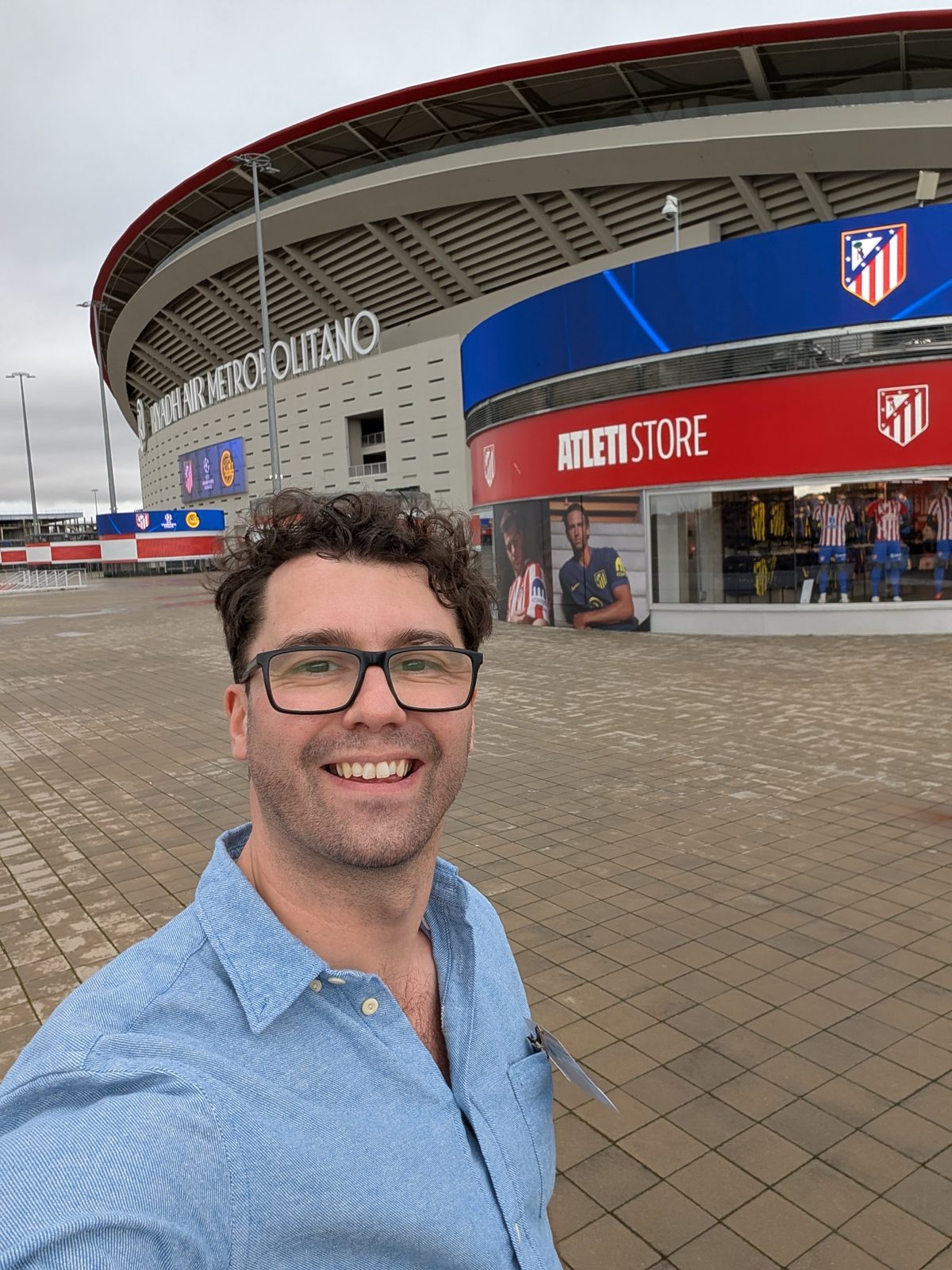 A selfie in front of the Riyadh Air Metropolitano stadium and the Atleti Store under a grey sky